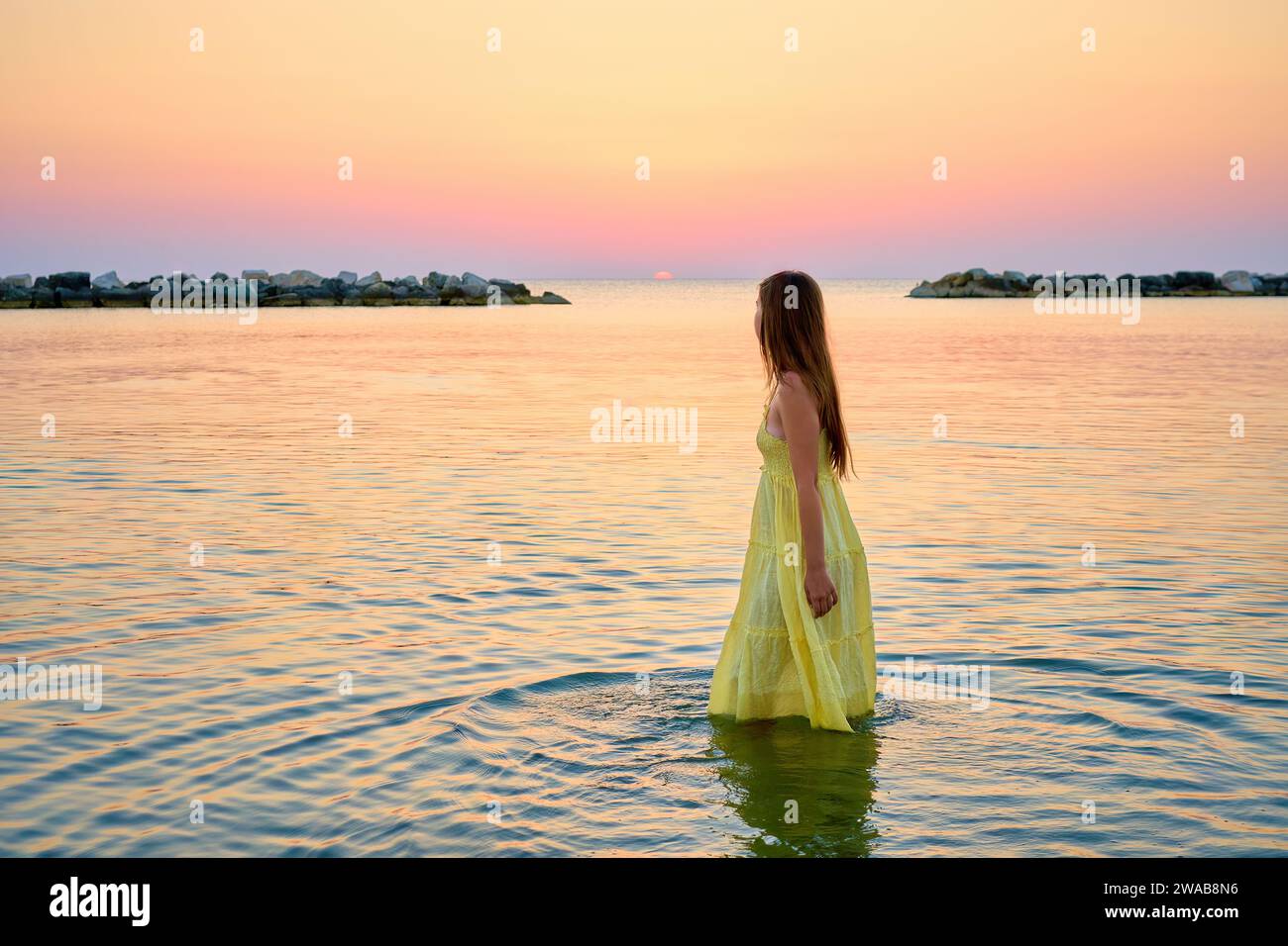 Una giovane donna con un vestito giallo brillante incontra l'alba, corre gioiosamente attraverso l'acqua di mare Foto Stock