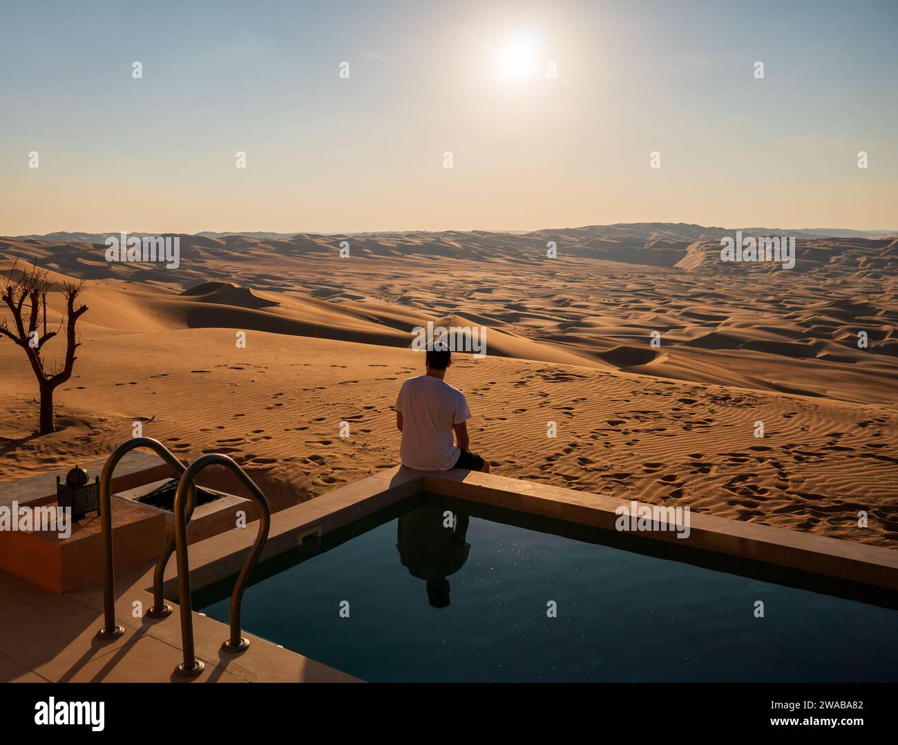 Un uomo seduto sul bordo di una piscina privata sullo sfondo di dune di sabbia e paesaggio desertico nel deserto di Rub'al Khali, Liwa. Sole brillante Foto Stock