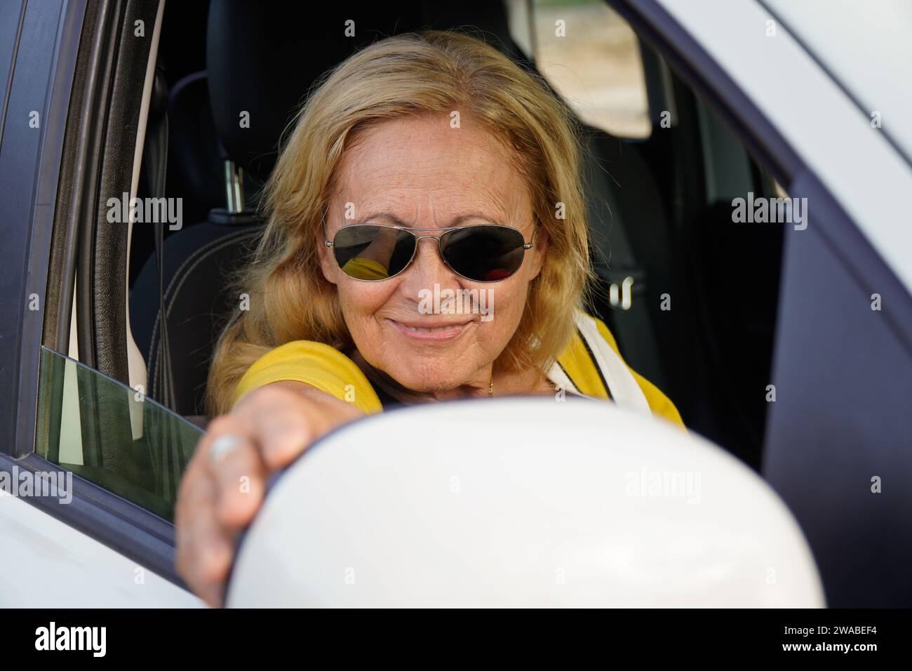 Donna matura con occhiali da sole e abiti estivi che regola lo specchietto retrovisore della vettura Foto Stock