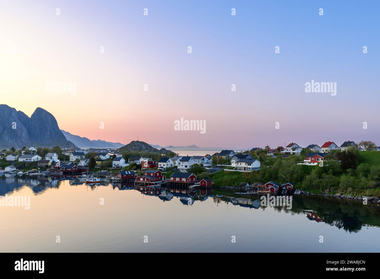 Le calme acque di Reine, le Isole Lofoten, sotto un cielo pastello, riflettono il pittoresco villaggio e il suo iconico rorbuer rosso, con aspre montagne Foto Stock