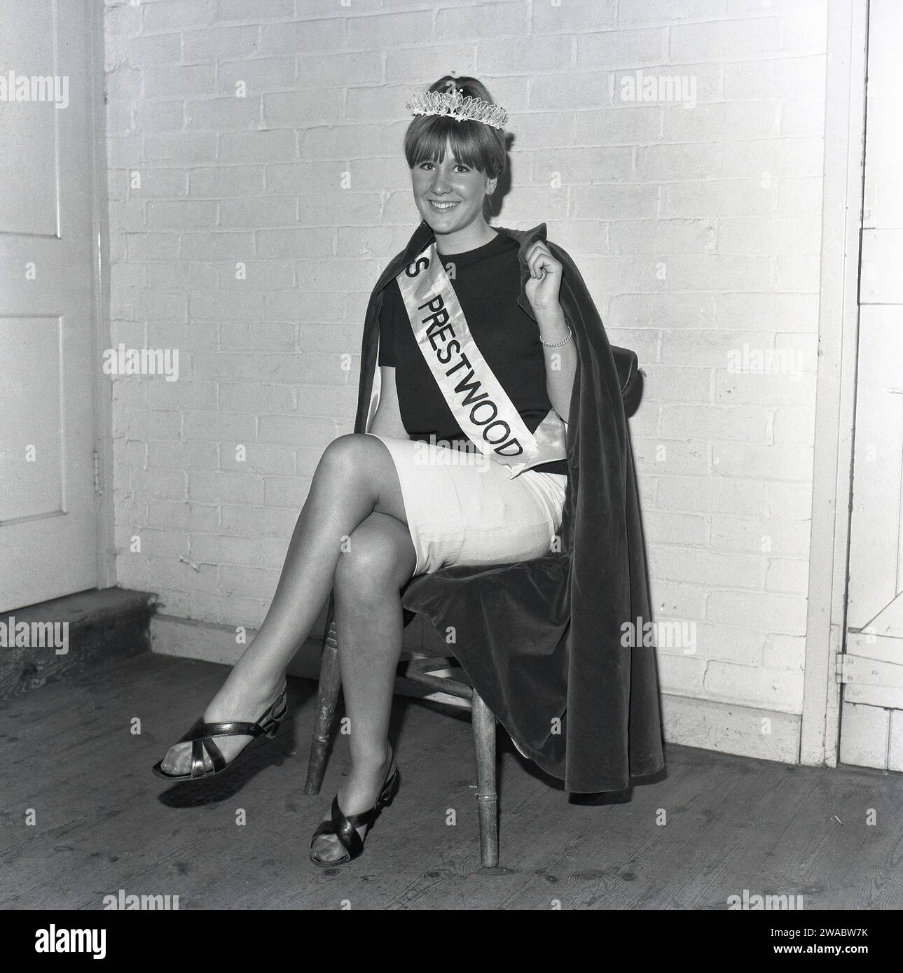 1964, storico, all'interno di un municipio del villaggio, una giovane donna seduta su una sedia con corona, fascia e mantello di velluto, incoronata "Miss Prestwood", la regina di bellezza del villaggio, Prestwood, Buckinghamshire, Inghilterra, Regno Unito. Foto Stock
