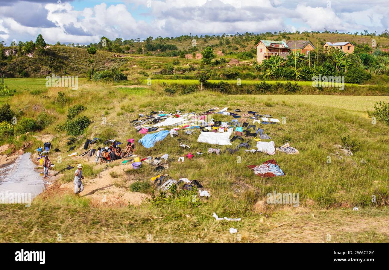 Behenjy, Madagascar - 25 aprile 2019: Gruppo di malgasci che fanno il bucato in un piccolo torrente, vestiti che si asciugano sull'erba vicino, cespugli verdi sul lato, sma Foto Stock