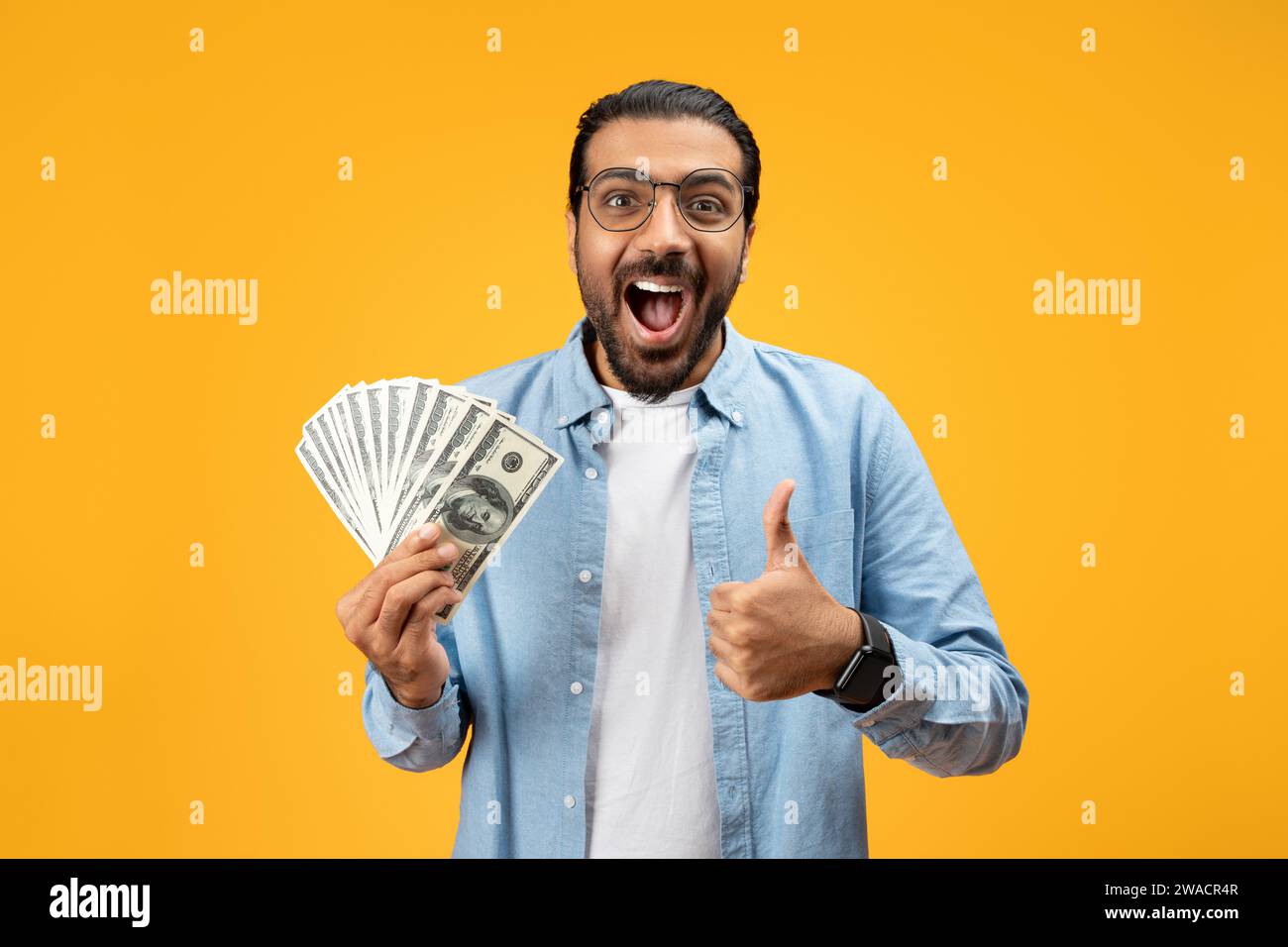 Un uomo estatico con una camicia in denim blu mostra un fan di banconote da 100 dollari mentre dà un pollice in su Foto Stock