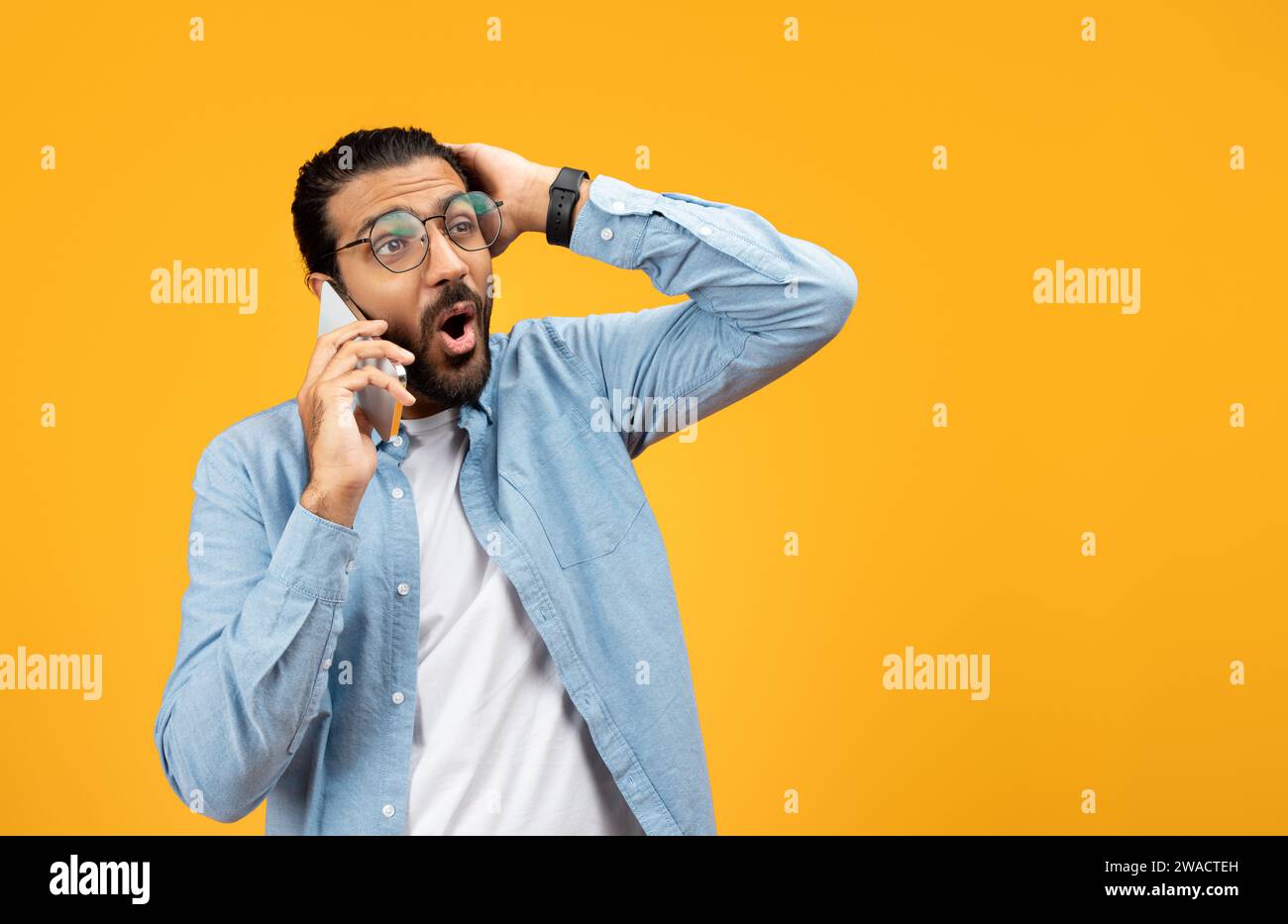 Uomo scioccato con una camicia blu in denim che parla al cellulare, che tiene la testa nell'incredulità o riceve Foto Stock