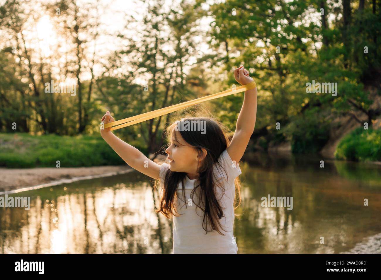 Adorabile bambina che solleva le braccia con una mini fascia sui polsi sopra la testa e allunga attrezzature sportive mentre si trova vicino al lago nella foresta. Junior a Foto Stock