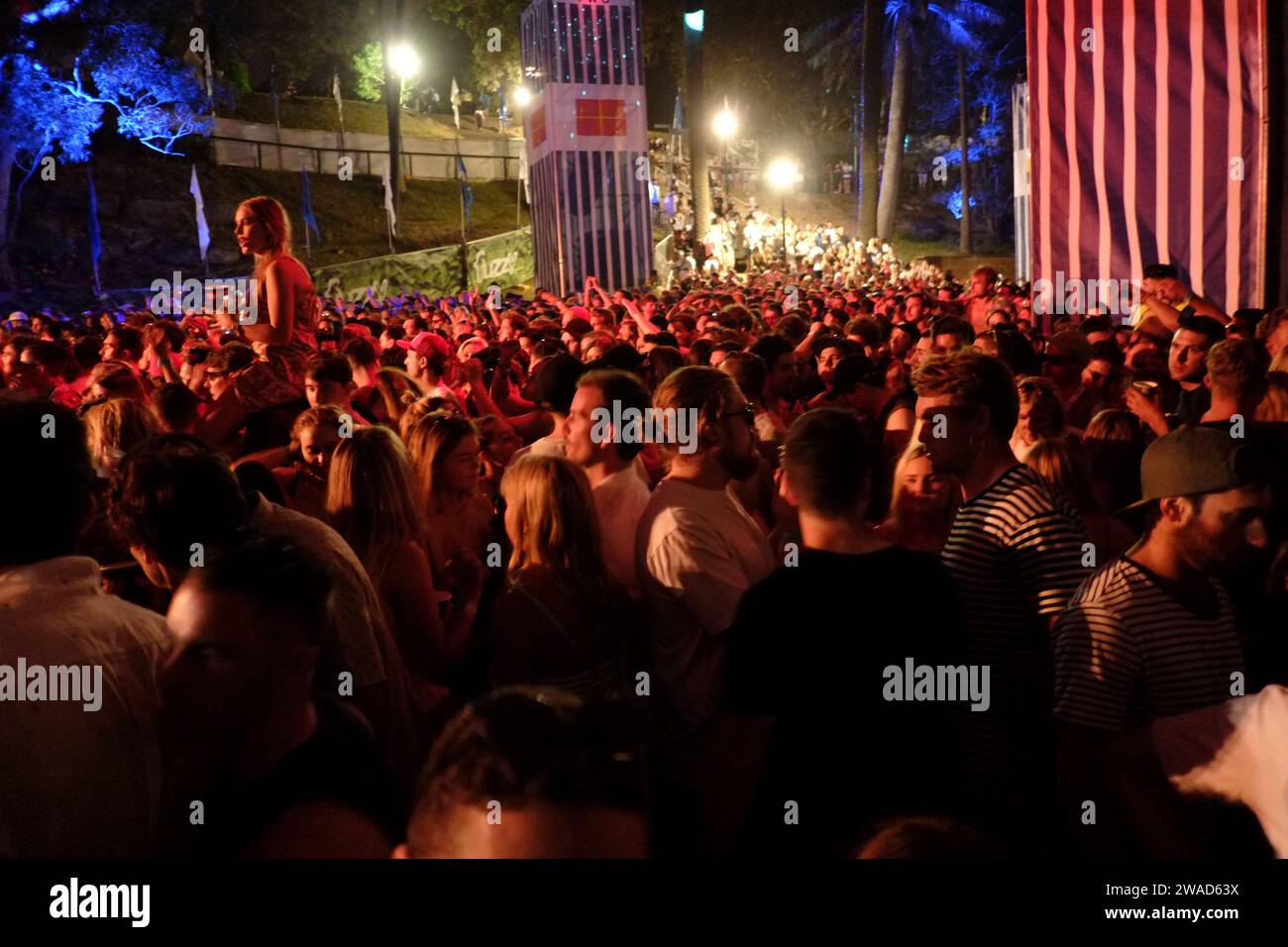Una folla ristretta di persone a una festa di musica all'aperto di notte, una donna che cavalca sulle spalle sopra il mare di persone che guardano il palco Foto Stock