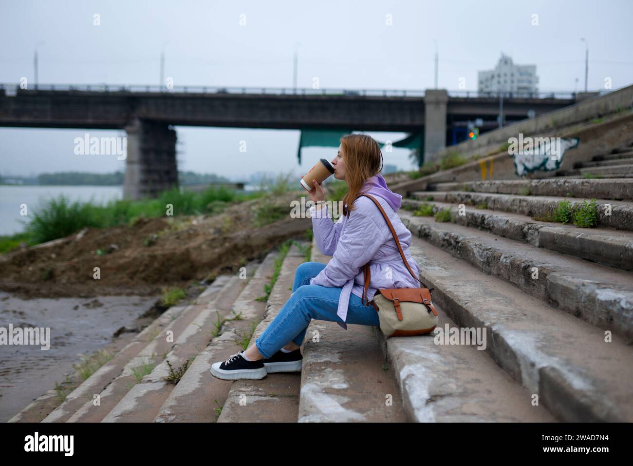 Donna premurosa seduta sui gradini e bevuta un caffè mentre guarda il fiume Foto Stock