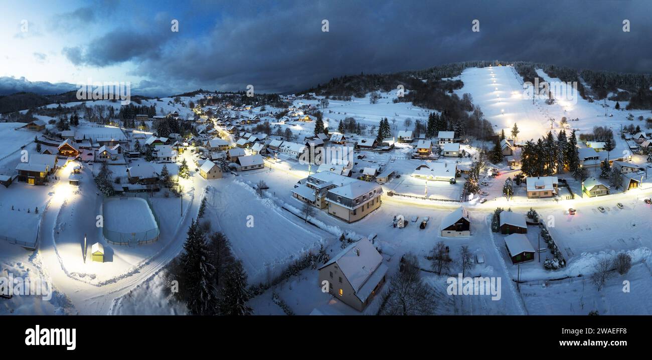 Vista della catena montuosa innevata e del villaggio con case illuminate situate in fondo alla collina di Slovkia al tramonto Foto Stock