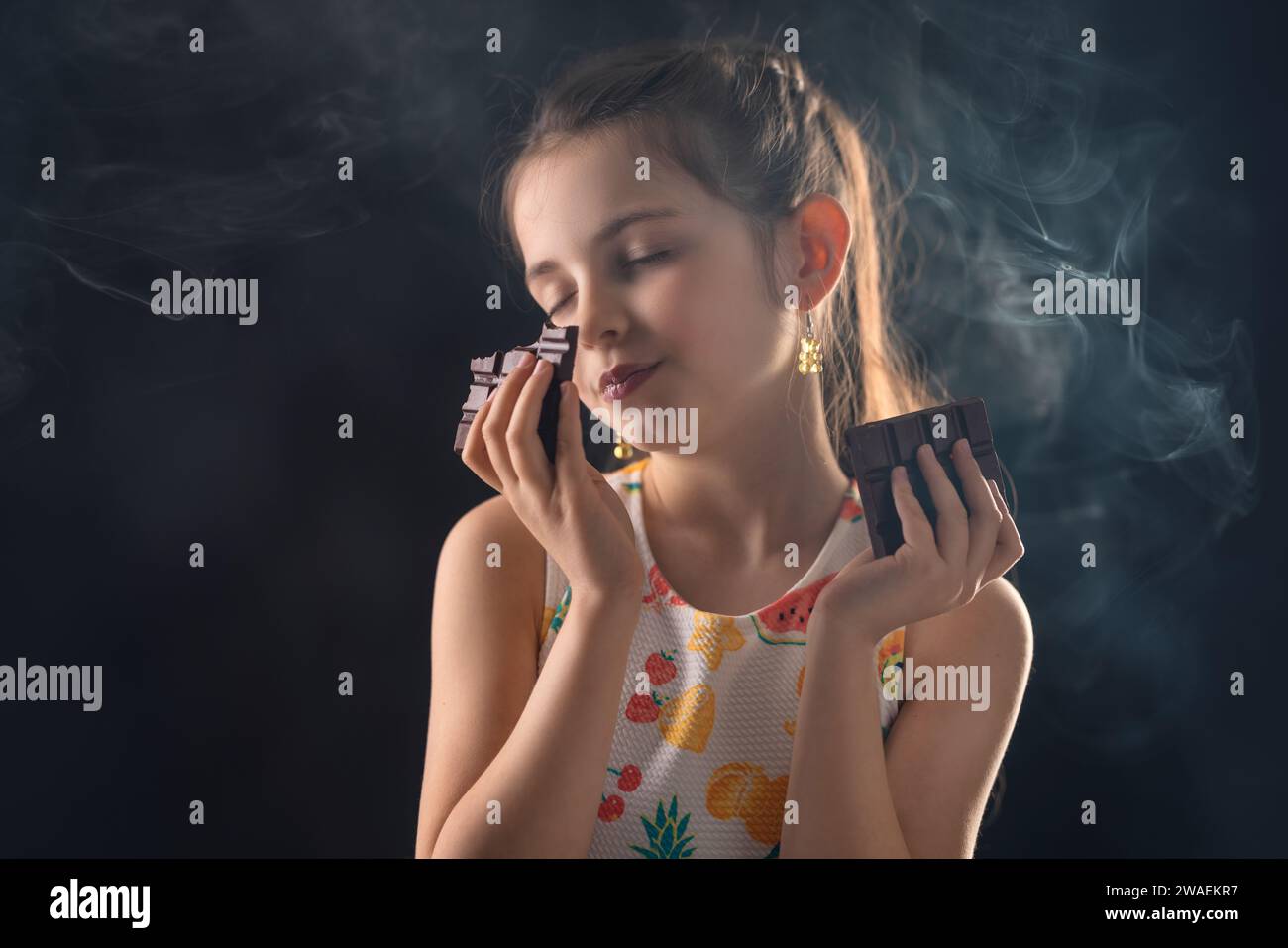 Una ragazza carina puzza e assaggia la barretta di cacao al cioccolato prima di mangiarla con nebbia e luce su sfondo nero Foto Stock
