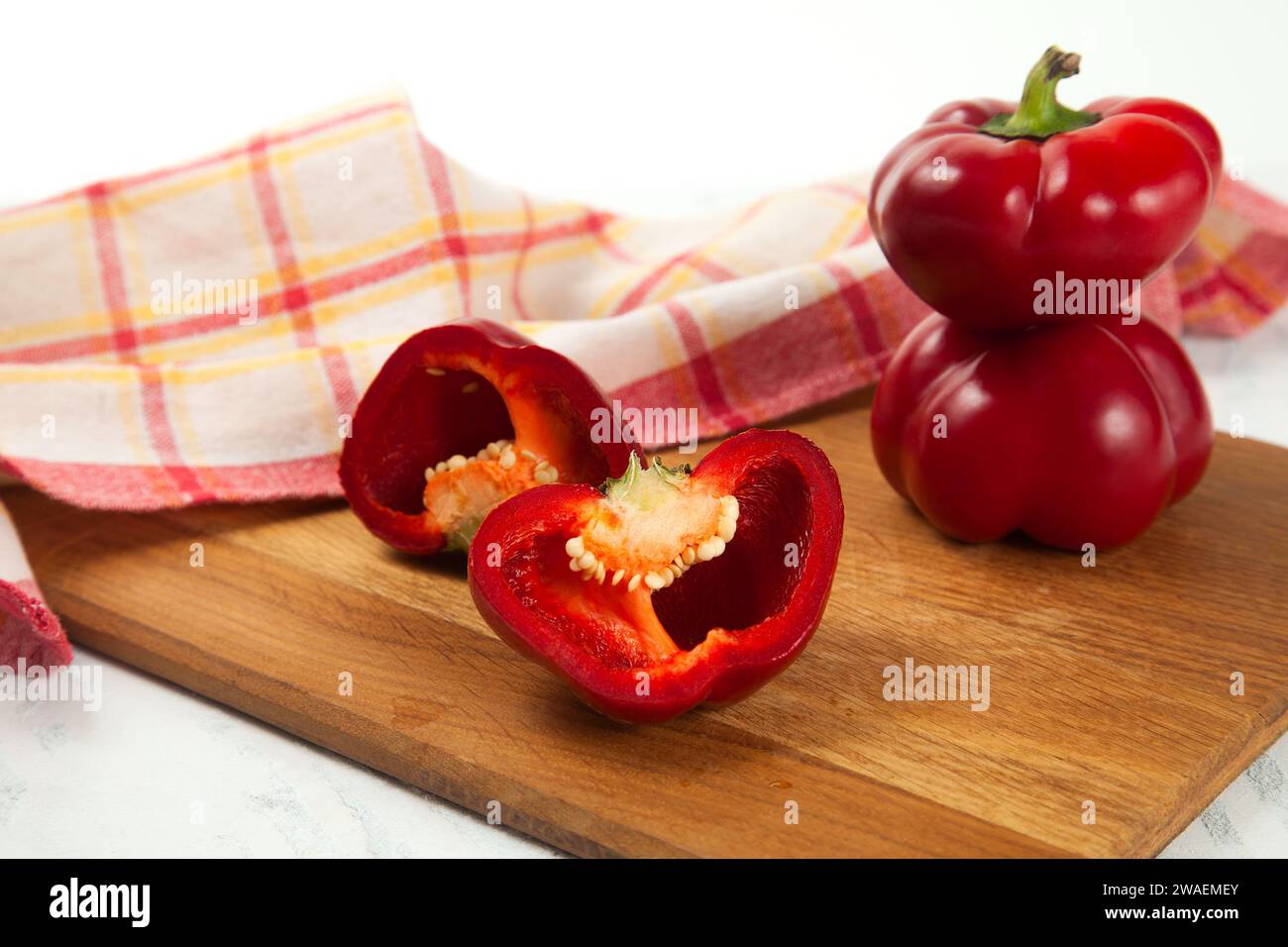 Tagliere con peperone rosso intero e mezzo (capsicum) noto come peperone dolce, paprika e asciugamano da cucina rosso su fondo di legno bianco. Foto Stock
