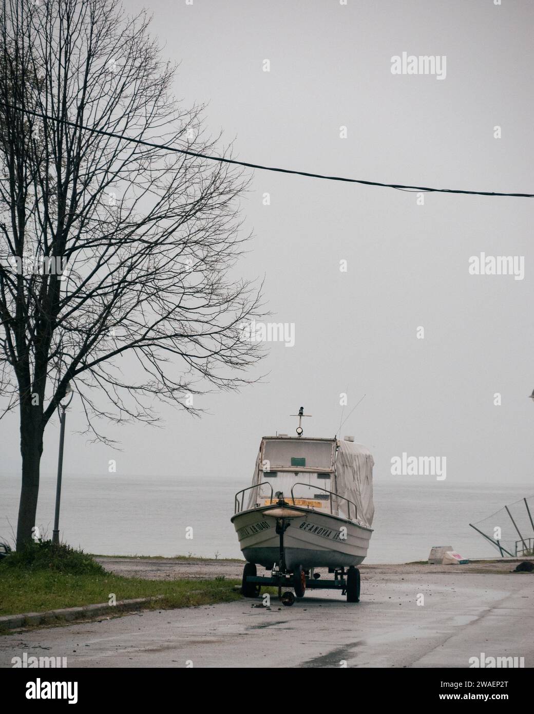 Piccola barca bianca sulla spiaggia in un villaggio greco in inverno accanto ad un albero senza foglie in Makedonia, Grecia Foto Stock