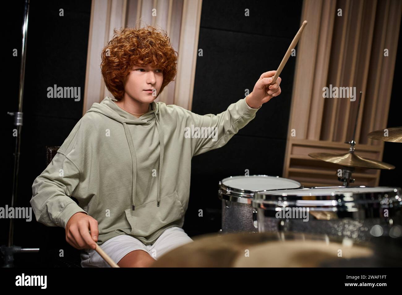 adorabile ragazzo adolescente concentrato con i capelli rossi in outfit di tutti i giorni che suona la batteria mentre è in studio Foto Stock