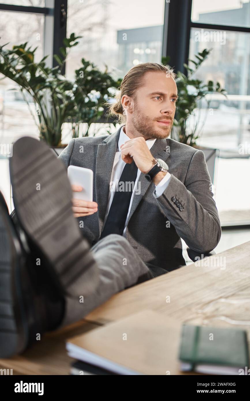 uomo d'affari rilassato e di successo in abito grigio seduto con le gambe sulla scrivania e guardando lontano in ufficio Foto Stock