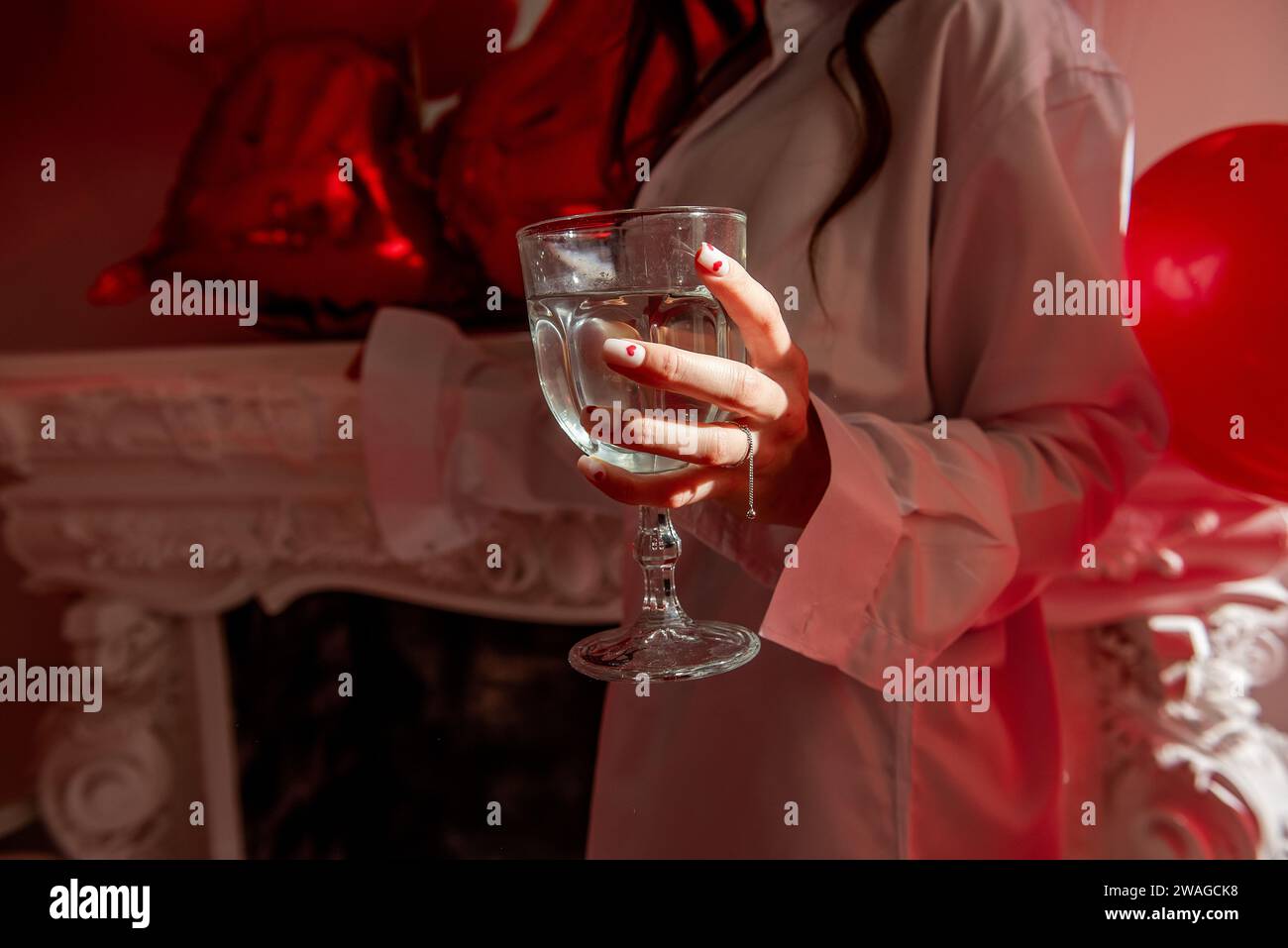 Primo piano della mano femminile con un bicchiere di champagne con manicure festive con cuori rossi sull'unghia, anello in argento. Festeggiamo San Valentino, fidanzamento Foto Stock