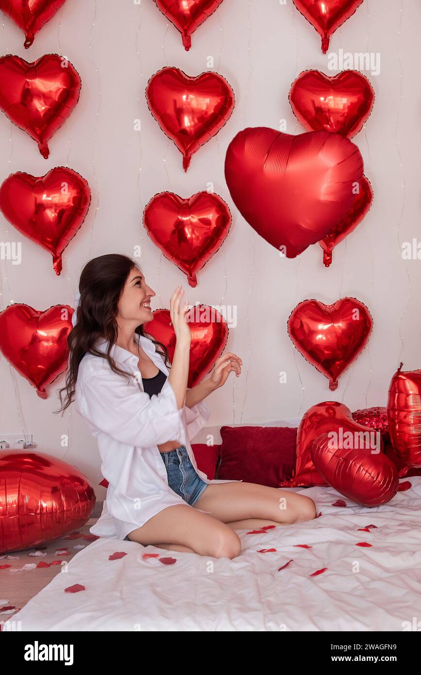 Bella donna che gioca con un palloncino a forma di cuore rosso su un letto adornato da lenzuola bianche petali di rosa, sorridente. Ragazza blogger che ingannano quasi creativo W Foto Stock