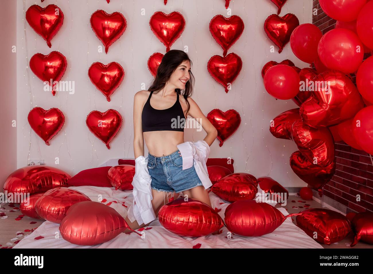 La giovane donna si inginocchia sul letto con petali di rosa, tra palloncini gonfiabili a forma di cuore. La ragazza blogger posa in pantaloncini in jeans corti in camicia bianca decorata Foto Stock