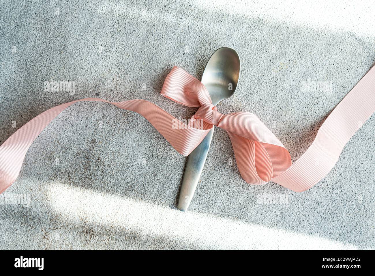 Un cucchiaio in argento legato a un delicato nastro rosa è adagiato su un panno grigio testurizzato, che presenta un dettaglio semplice ma elegante Foto Stock