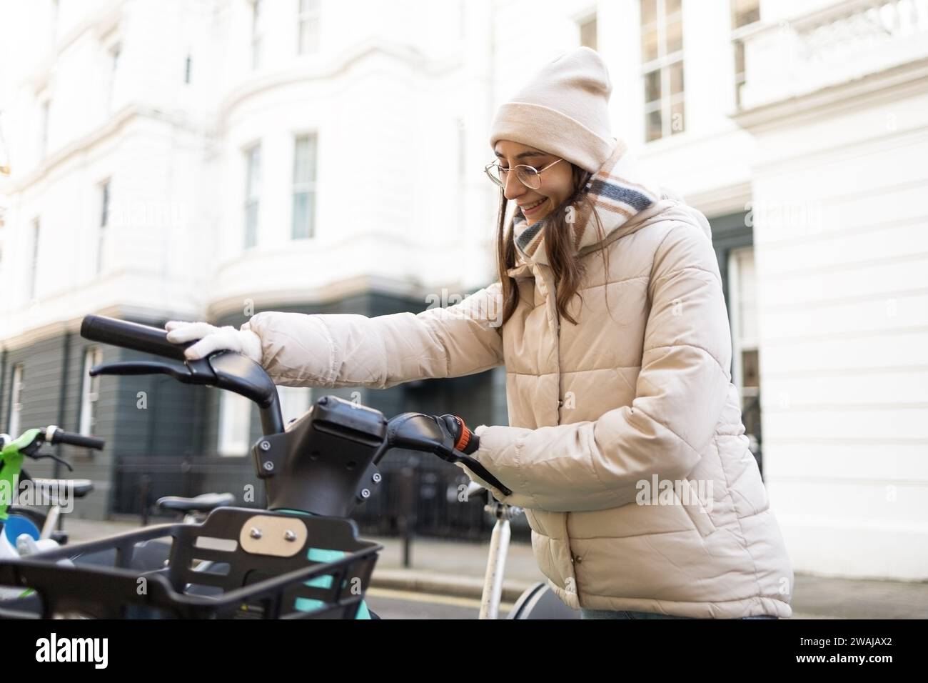 Vista laterale di una giovane donna positiva con una giacca calda e un berretto in piedi accanto a uno scooter elettrico sulla strada della città, davanti alla facciata di un moderno appartamento bianco di giorno Foto Stock