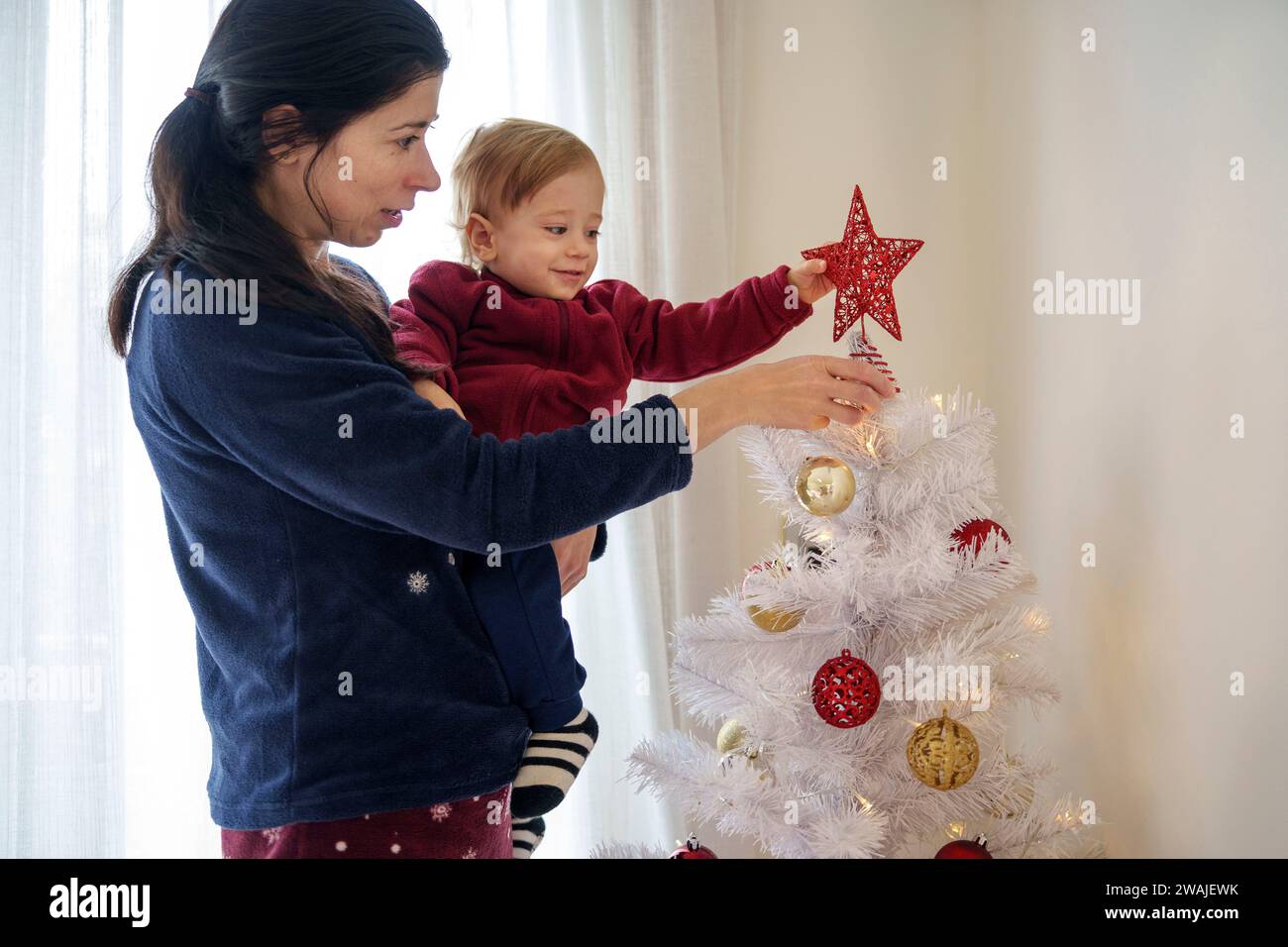 Bambino di 16 mesi che aiuta sua madre a preparare le decorazioni natalizie sull'albero di Natale Foto Stock