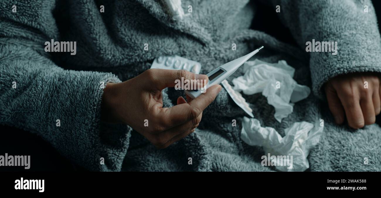 un uomo a casa, che indossa un soffice abito da casa grigio, circondato da tessuti usati, sta per misurare la sua temperatura con un termometro, in forma panoramica Foto Stock
