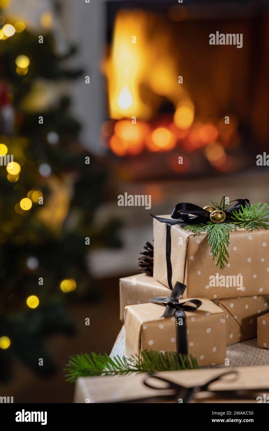Regali di Natale avvolti con fiocchi a nastro nero sullo sfondo di un camino in fiamme e un albero di natale decorato con luci. Calda atmosfera invernale per famiglie Foto Stock