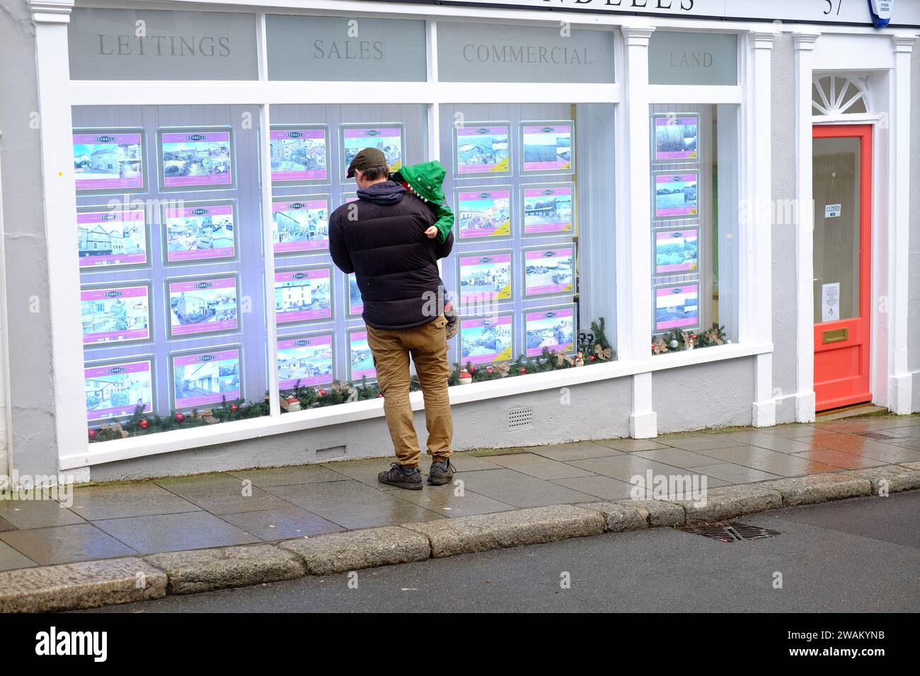 L'uomo che tiene in braccio un bambino si trova fuori dall'ufficio dell'agente immobiliare e guarda gli annunci di vendita e affitto di immobili nella finestra. Totnes, Devon, Regno Unito Foto Stock