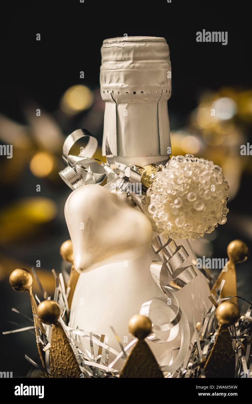 Piccola bottiglia bianca con vino frizzante in una corona metallica dorata decorata da un cuore bianco. Primo piano verticale. Foto Stock
