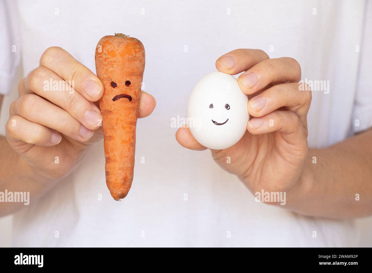 Una carota con un sorriso triste e un uovo con un sorriso gioioso nelle mani di una ragazza su sfondo bianco, tristezza, tristezza, prodotti alimentari e dieta Foto Stock