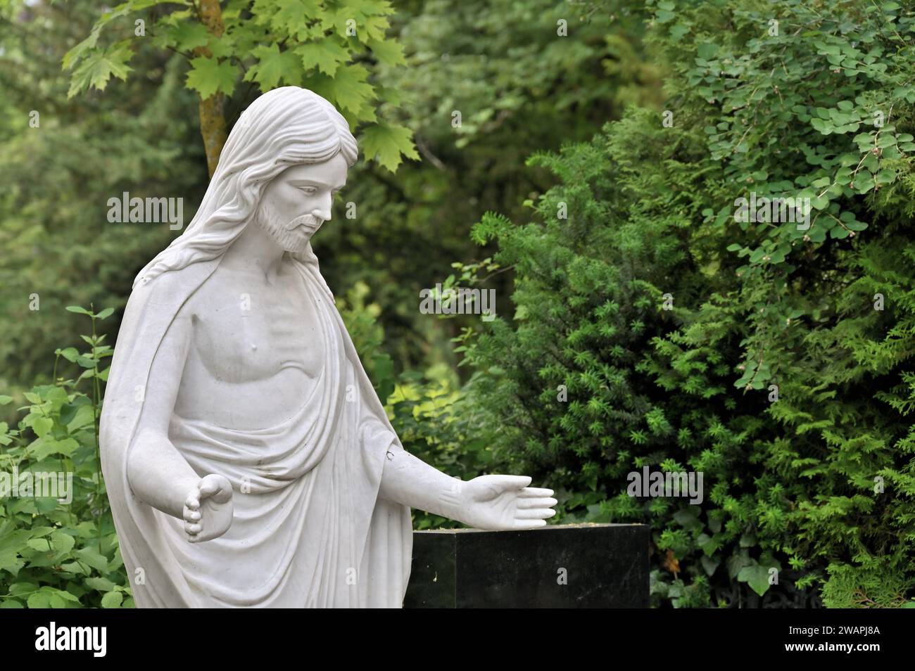 Statua bianca di Cristo con le mani tese di fronte al verde fogliame Foto Stock