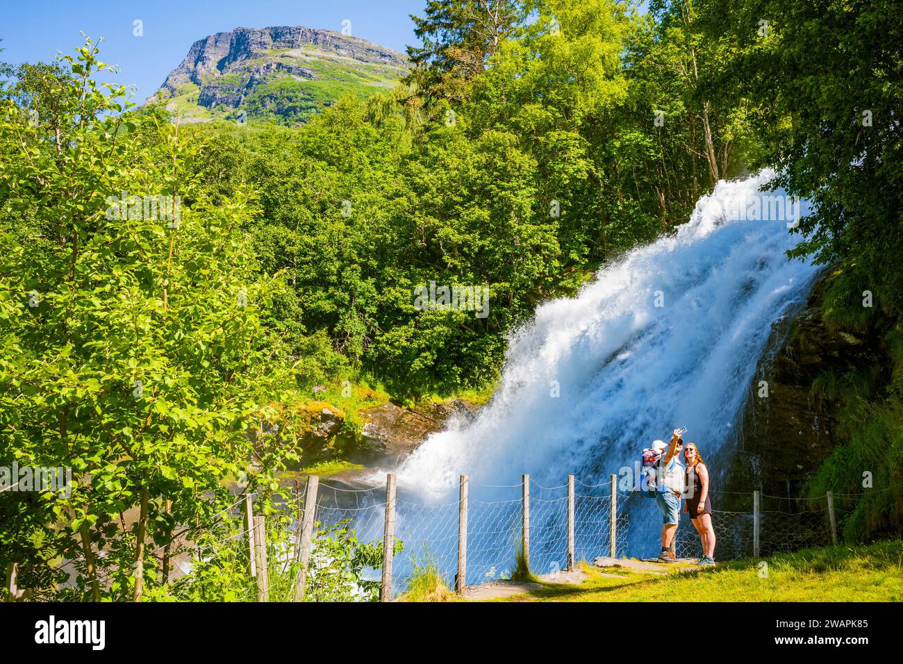 Geiranger, Norvegia, 26 giugno 2023: Una giovane famiglia scatta foto vicino a una piccola cascata chiamata Kleivafossen situata in un campeggio nel piccolo villaggio a Foto Stock