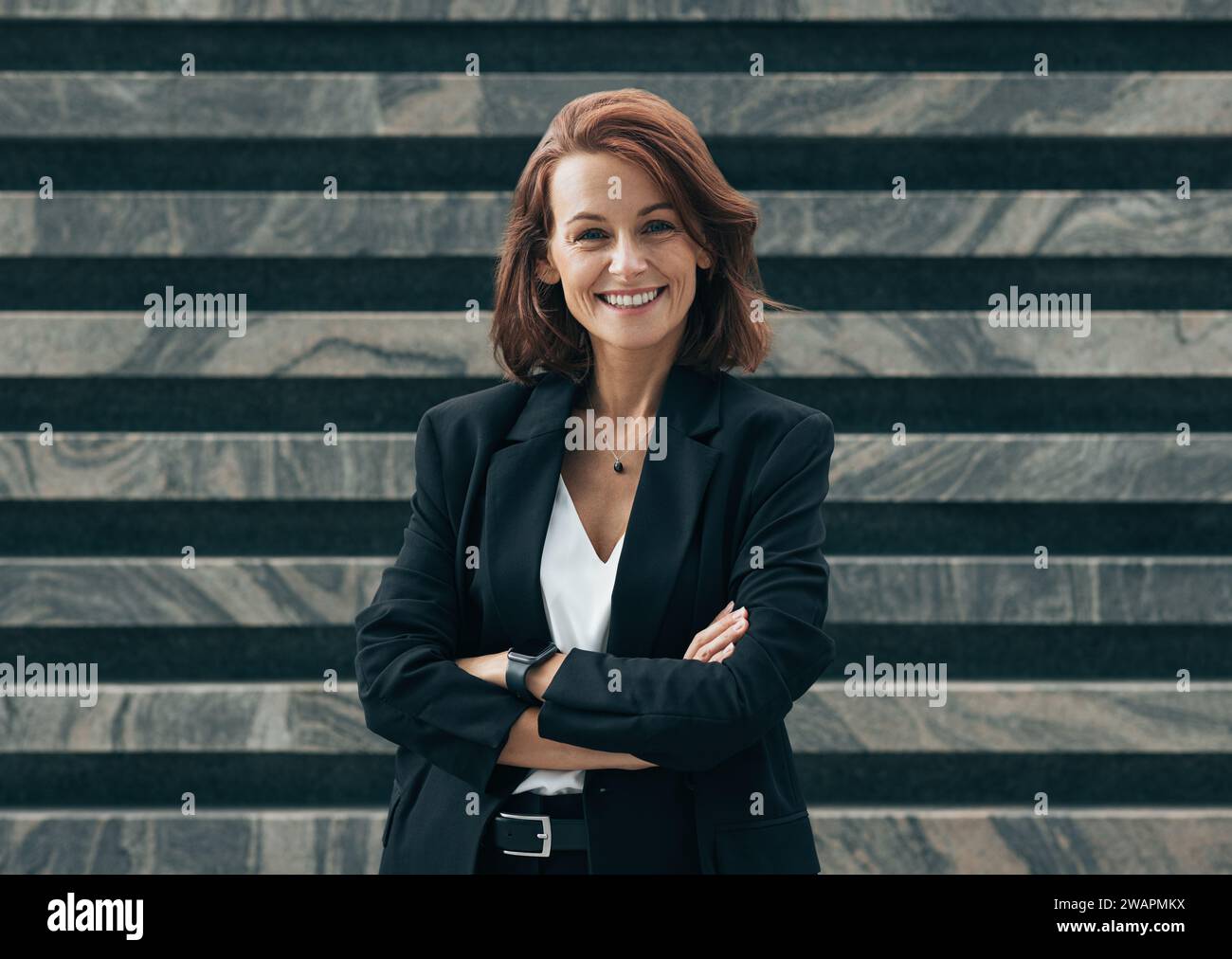 Donna felice con capelli di zenzero in abbigliamento formale che guarda la fotocamera. Giovane donna sicura di sé di mezza età con le braccia incrociate in piedi all'aperto e sorridente. Foto Stock