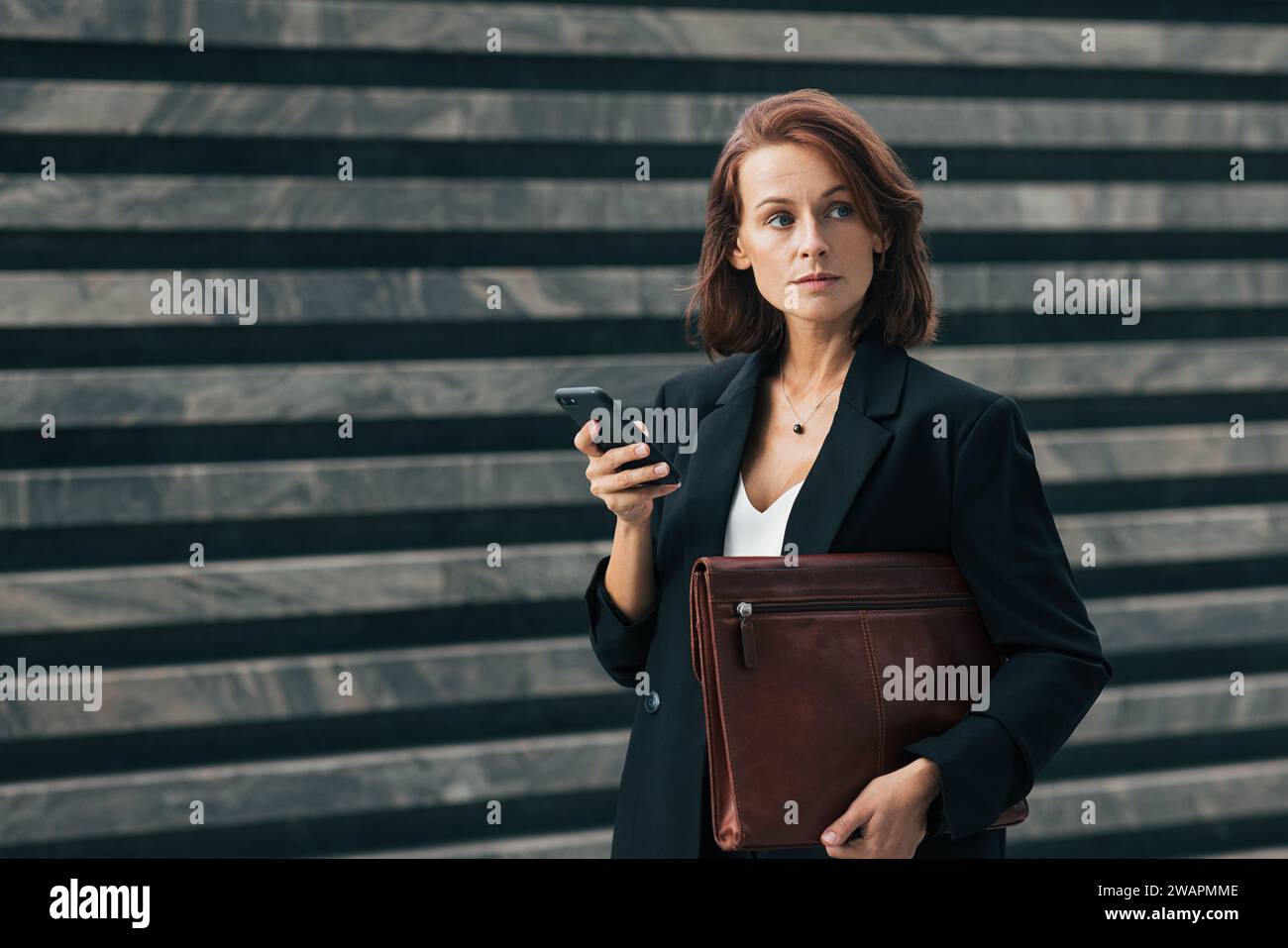 Donna d'affari sicura di sé con i capelli di zenzero. Donna di mezza età che tiene lo smartphone e guarda lontano. Donna in abiti formali che reggono una piega in pelle Foto Stock