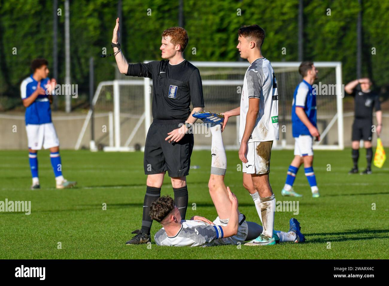 Landore, Swansea, Galles. 6 gennaio 2024. L'arbitro di partita Ollie Taylor si ferma mentre Iestyn Jones di Swansea City aiuta il compagno di squadra Jack Fanning durante la partita Under 18 della Professional Development League tra Swansea City e Ipswich Town alla Swansea City Academy di Landore, Swansea, Galles, Regno Unito il 6 gennaio 2024. Crediti: Duncan Thomas/Majestic Media/Alamy Live News. Foto Stock