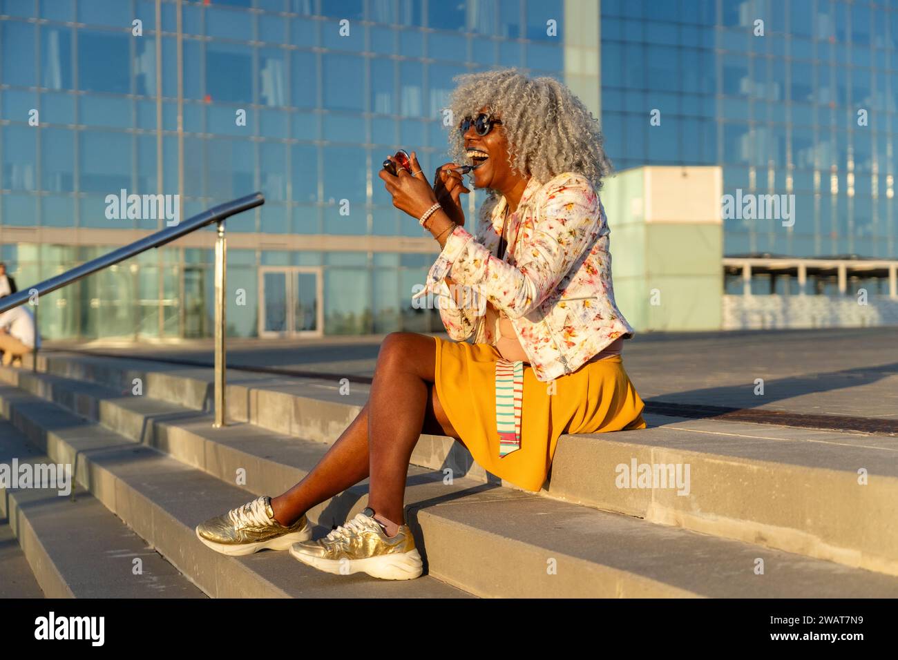 Signora nera espressiva con capelli afro e occhiali da sole che indossano rossetto blu nel soleggiato centro della città. Concetto: pro-aging, fiducia in se stessi Foto Stock