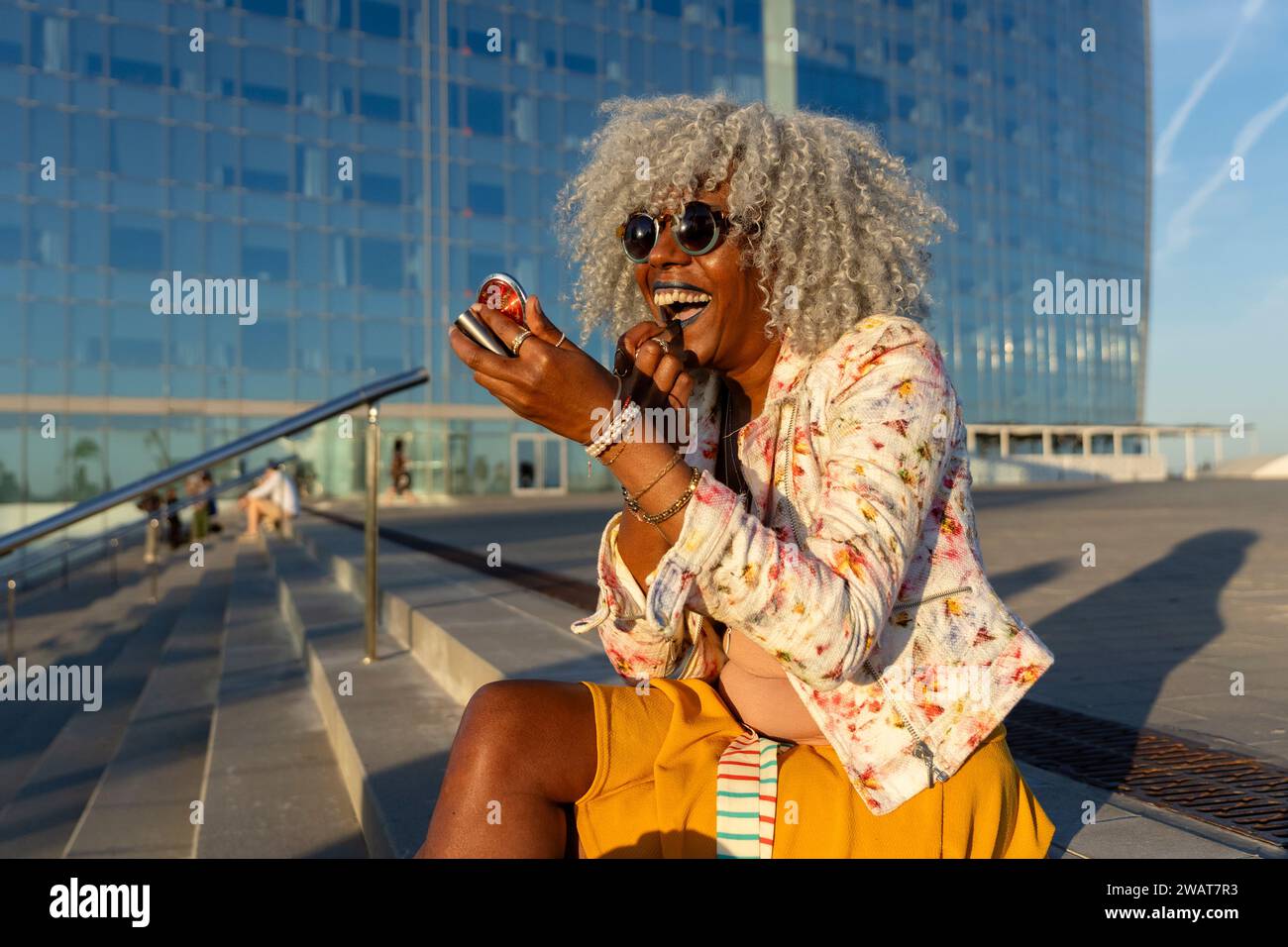 Signora nera espressiva con capelli afro e occhiali da sole che indossano rossetto blu nel soleggiato centro della città. Concetto: pro-aging, fiducia in se stessi Foto Stock