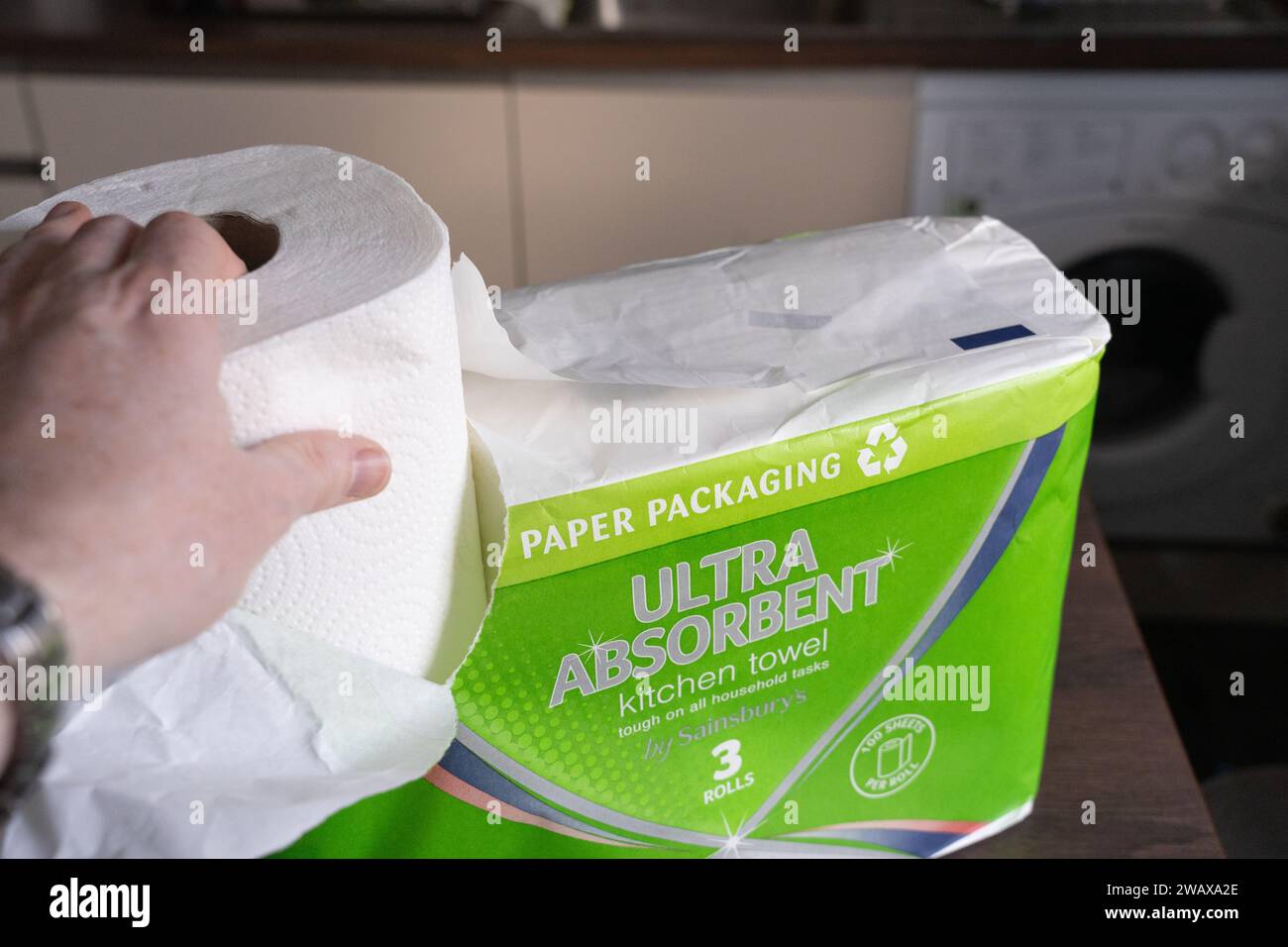 Una mano che rimuove un rotolo di asciugamani da cucina del marchio Sainsbury con imballaggio di carta in una cucina, Regno Unito. Concetto: Eliminazione degli sprechi di plastica, assenza di plastica Foto Stock