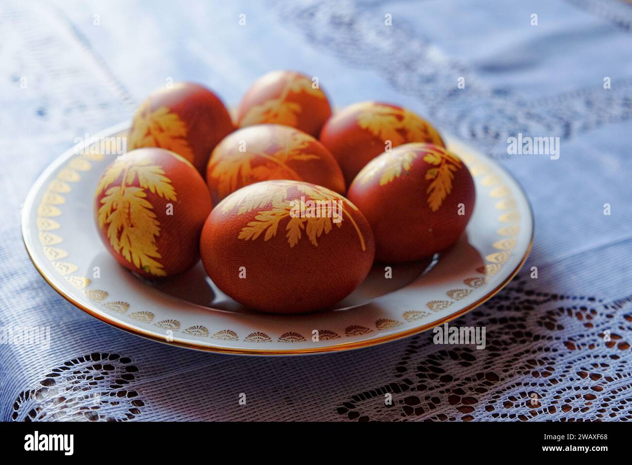L'immagine mostra sette uova marroni con motivi a foglia gialla su un piatto bianco con bordo dorato, adagiato su una tovaglia di pizzo blu e bianco. Foto Stock