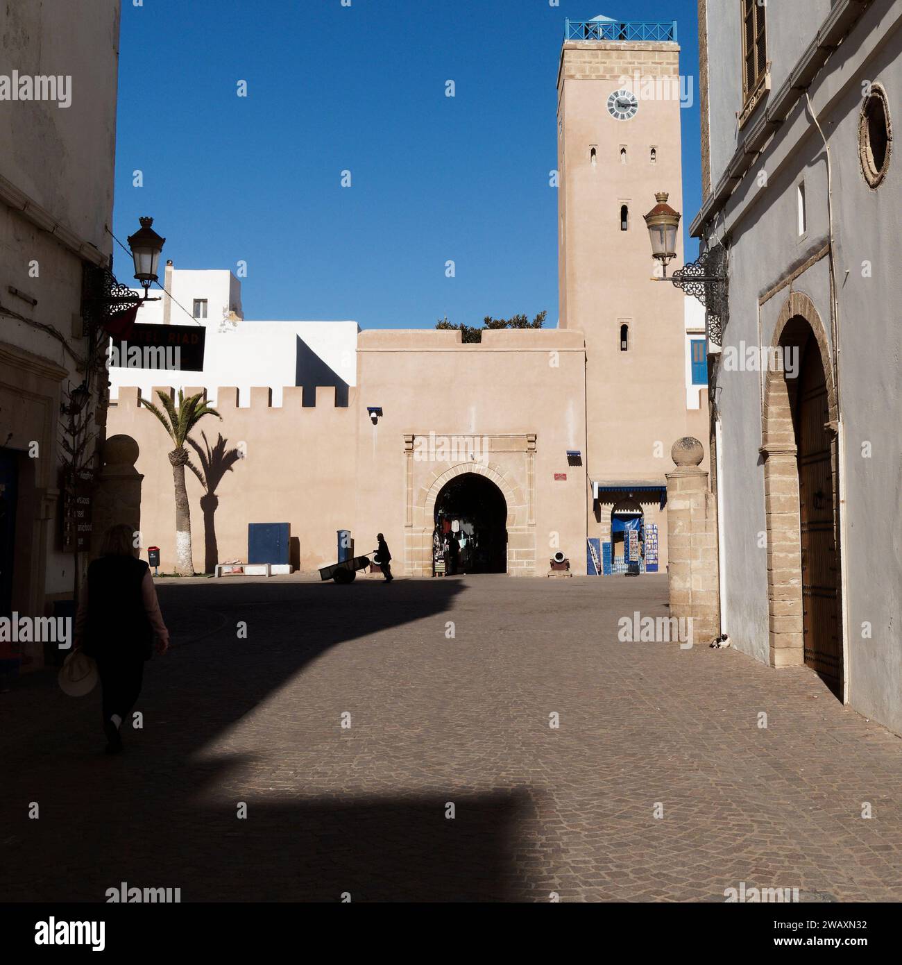 Strada nella storica medina con la torre dell'orologio davanti mentre qualcuno in silhouette spinge un carro nella città di Essaouira, in Marocco. 7 gennaio 2024 Foto Stock