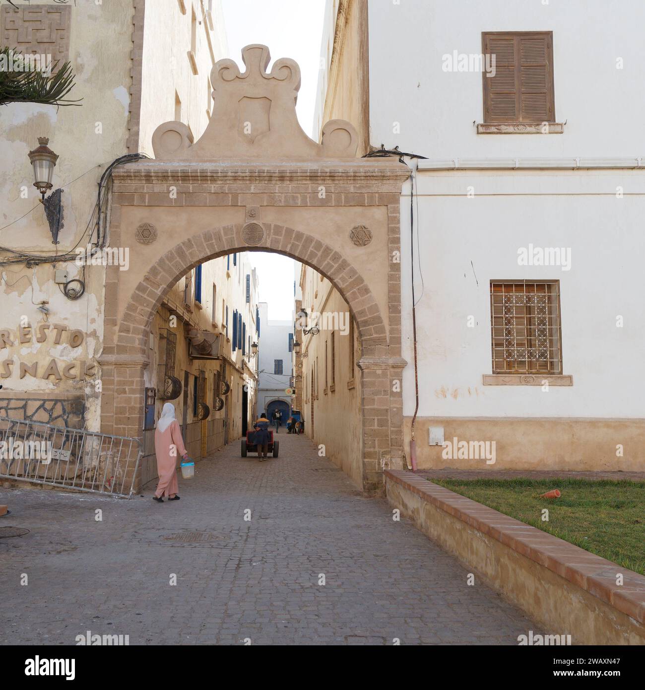 Una donna del posto in abiti tradizionali cammina verso un cancello storico con l'uomo che spinge un carro nella medina storica. Essaouira, Marocco. 7 gennaio 2024 Foto Stock