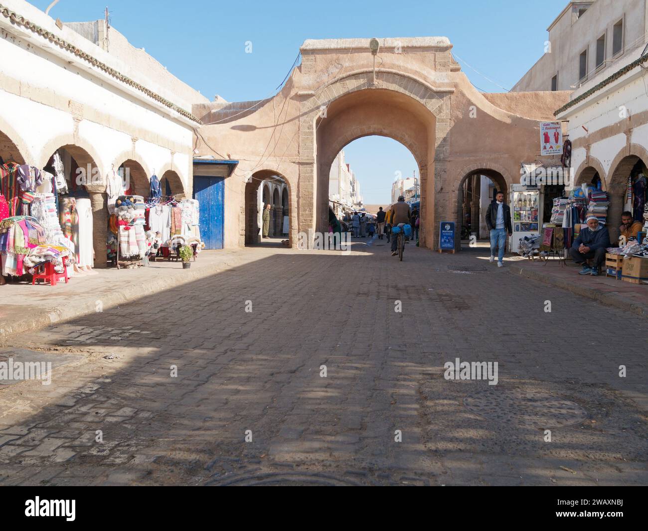 Strada principale con arco e negozio di abbigliamento mentre un uomo va in bicicletta nella città di Essaouira, in Marocco. 7 gennaio 2024 Foto Stock