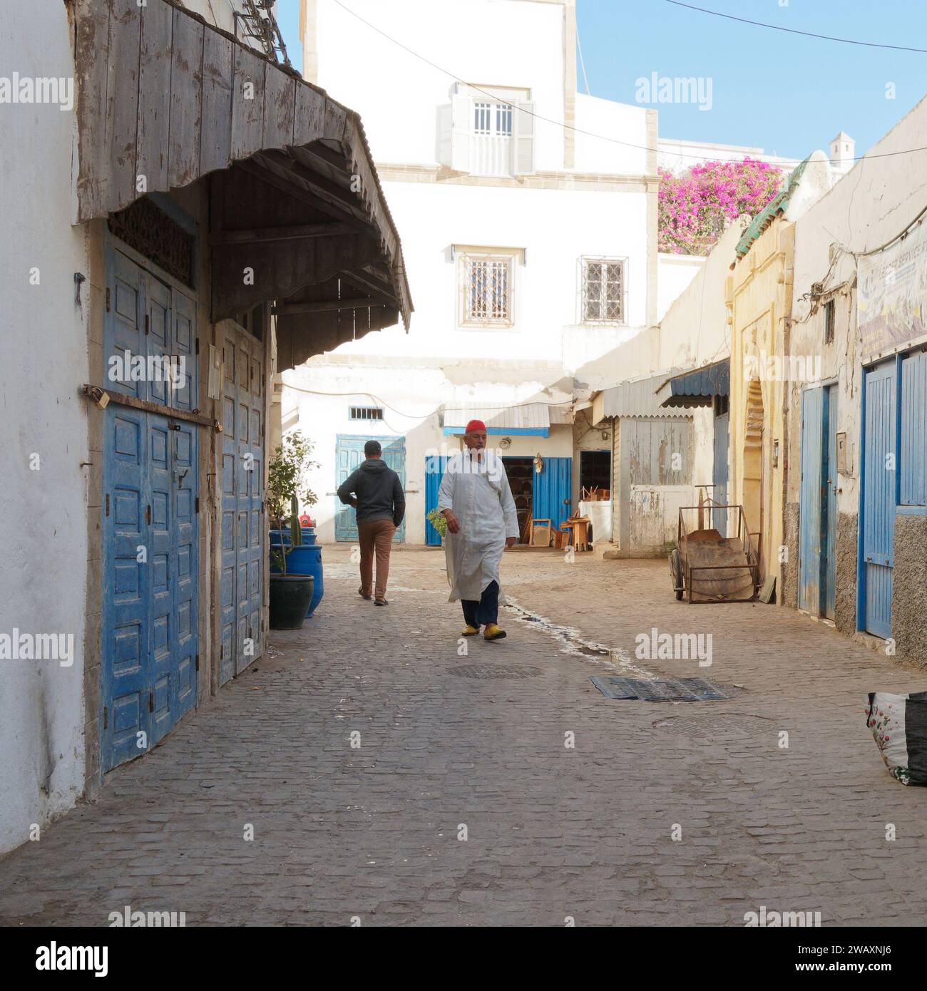 Un uomo del posto con cappello rosso cammina lungo una strada con porte blu nella storica medina della città di Essaouira, in Marocco. 7 gennaio 2024 Foto Stock