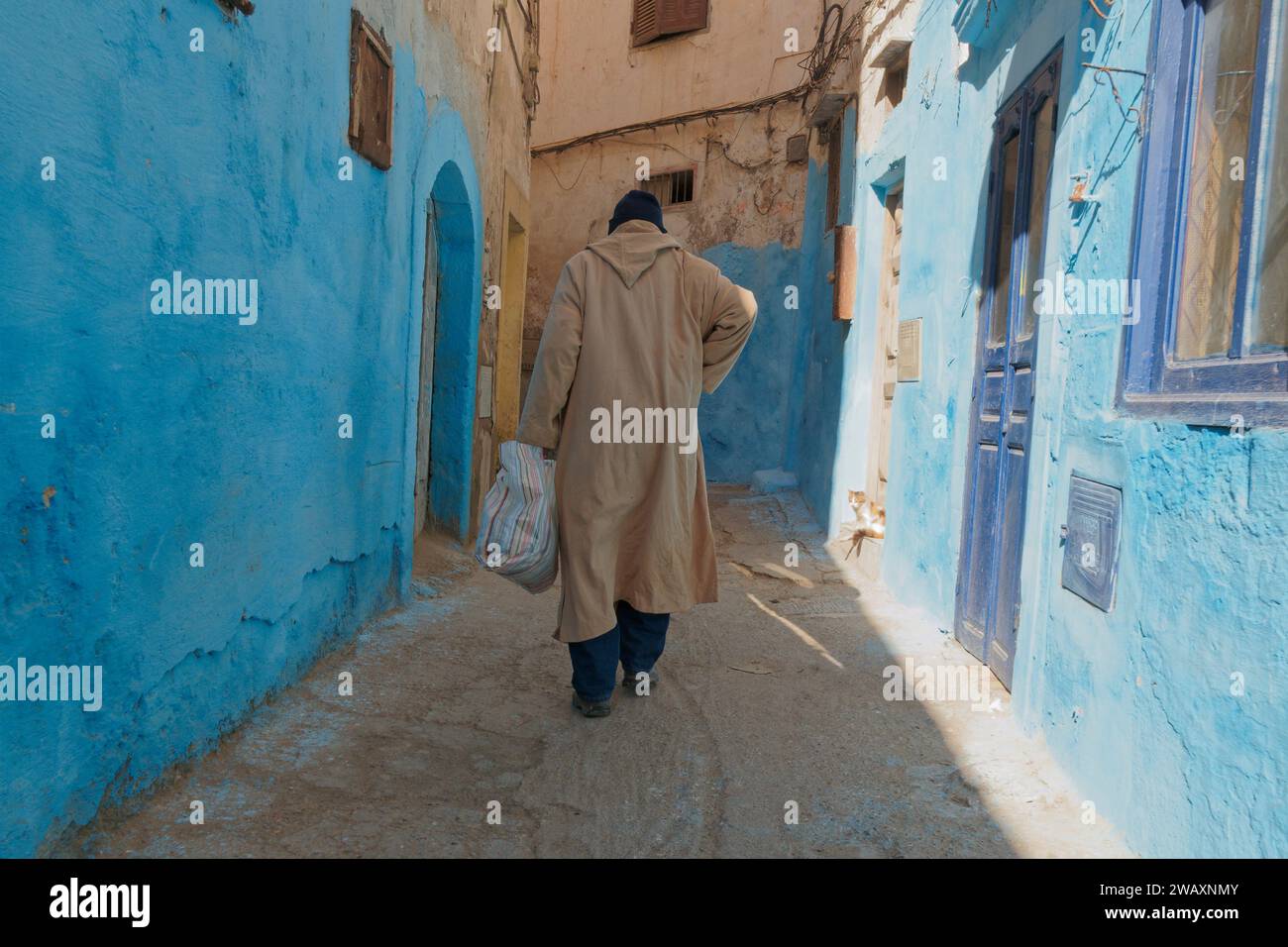 L'uomo locale cammina attraverso una strada stretta e colorata con mura blu nella storica medina nella città di Essaouira, in Marocco. 7 gennaio 2024 Foto Stock