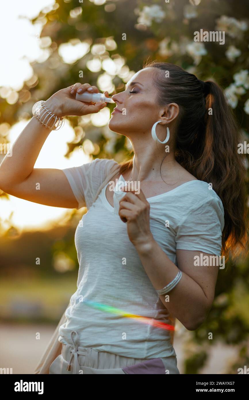 Ora legale. donna alla moda con camicia bianca che utilizza spray nasale vicino all'albero fiorito. Foto Stock