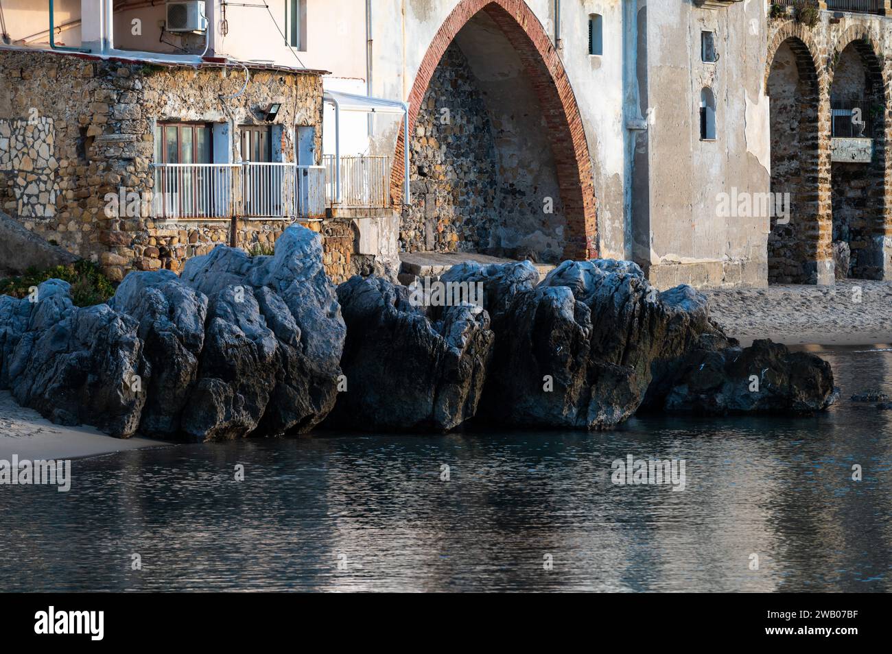 Cefalù, Sicilia, Italia, 19 dicembre 2023 - il mare, le rocce e un dettaglio di un edificio storico ad arco sul lungomare Foto Stock