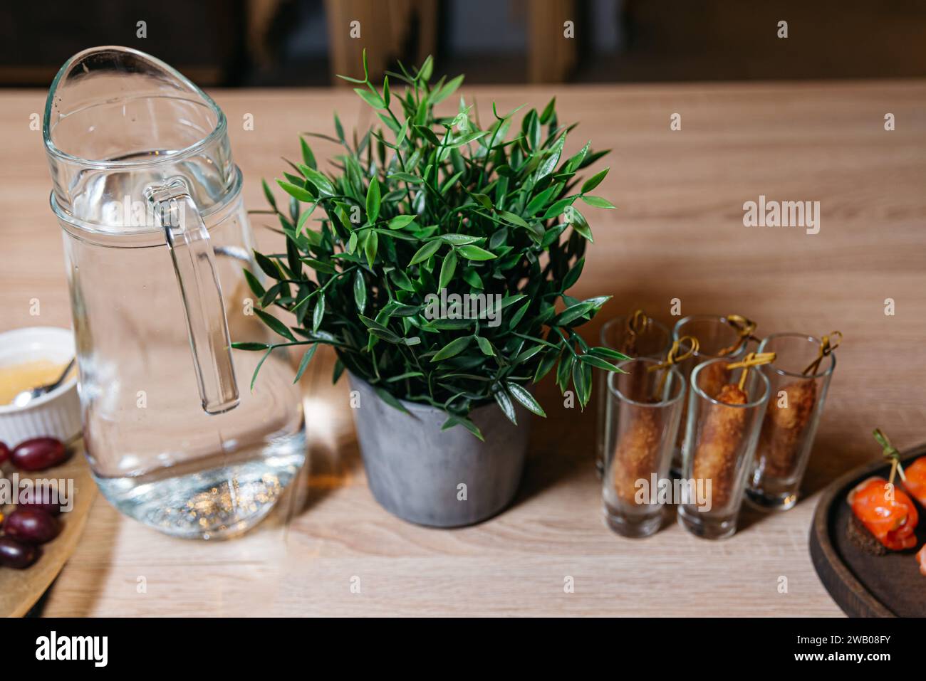 Una caraffa d'acqua contemporanea in vetro accanto a una lussureggiante pianta in vaso verde su un tavolo di legno, creando un ambiente pulito e rinfrescante. Foto Stock