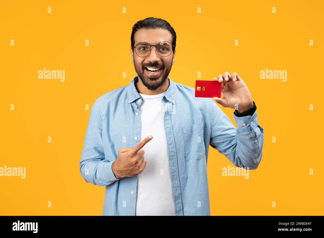Un uomo allegro con una camicia in denim che regge una carta di credito rossa e la punta con l'altra mano Foto Stock