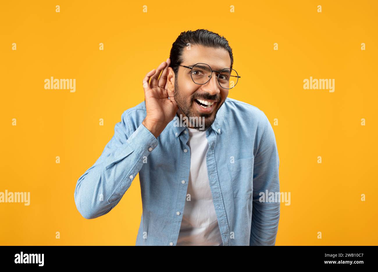 Un uomo delizioso con una camicia in denim blu con occhiali che avvolgono la mano vicino all'orecchio in un gesto di ascolto Foto Stock