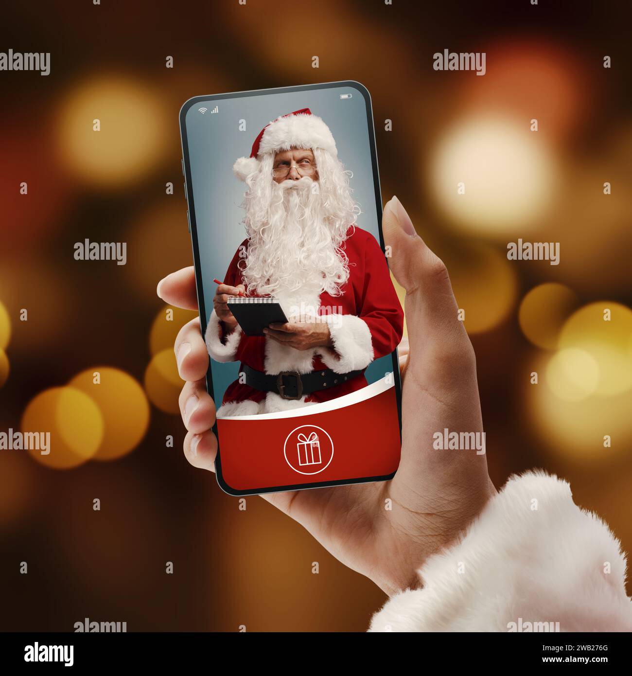 Mano che regge uno smartphone con Babbo Natale che scrive una lista dei desideri su un blocco note Foto Stock