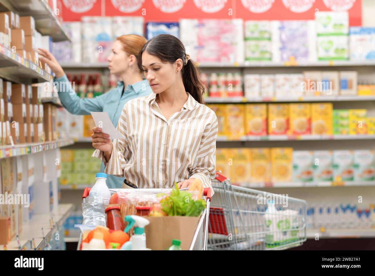 Giovane donna pensiva con carrello pieno al supermercato, sta controllando la sua lista di generi alimentari e il suo concetto di spesa Foto Stock