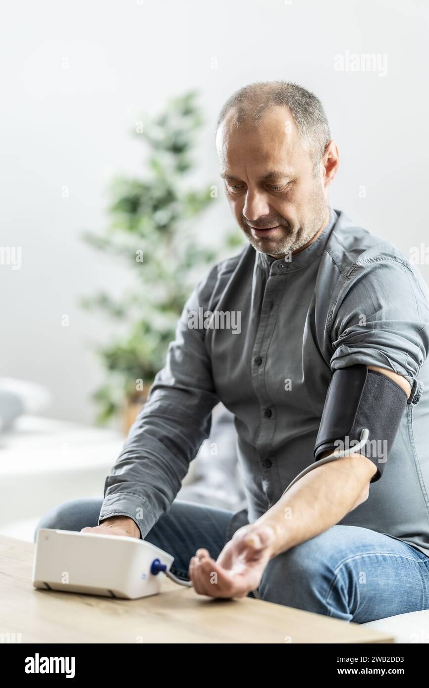 Un uomo maturo che controlla la pressione con un misuratore digitale della pressione a casa. Foto Stock